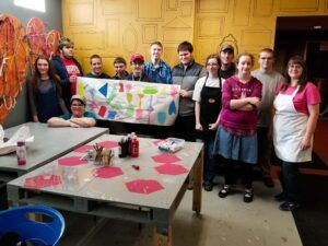 a large group of teenagers stand in front of a worktable holding up a paper that they have all added art to. Everyone is smiling
