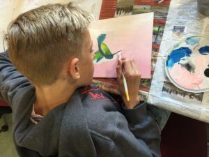 from an over-the-shoulder point of view, a young man is painting the beak on a hummingbird painting