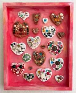 a mixed media piece with several ceramic appearing hearts with tiles, glass gems, and assorted doodads pressed into them, all arranged in a large square box painted a light red