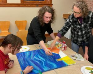 a teacher in plaid points at a painting that is being worked on by two students of work of art who are painting them.