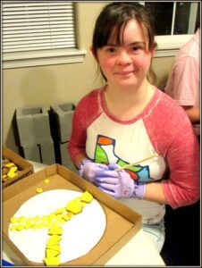 A girl sits looking at the camera holding a brush, in front of her is an artwork made of mixed media in the shape of the letter 'Y'