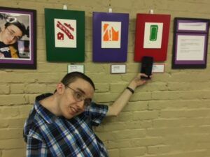 A student points to his artwork - several different items are hung on the wall behind him