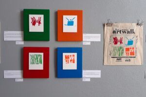 prints hung on a wall, text describing the artist next to each print, and a tote bag with the same prints hung next to them, with the artist's signatures on the tote bag.